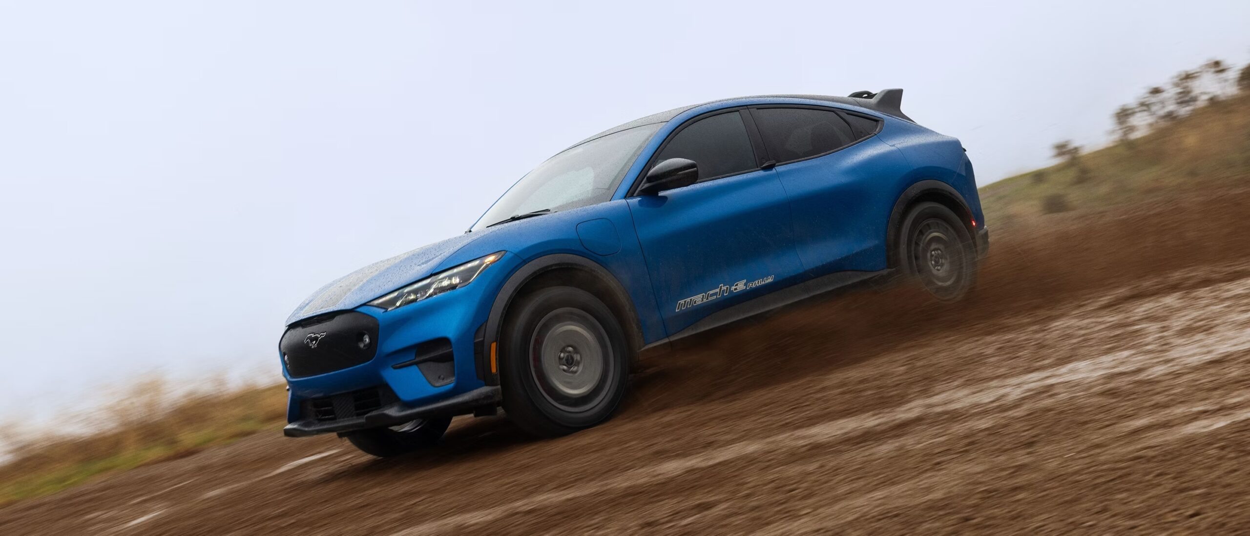 A blue Ford Mustang Mach-E Rally driving fast on a dirt track, kicking up a cloud of dust. The car is shown at a dynamic angle against a muted, overcast background.